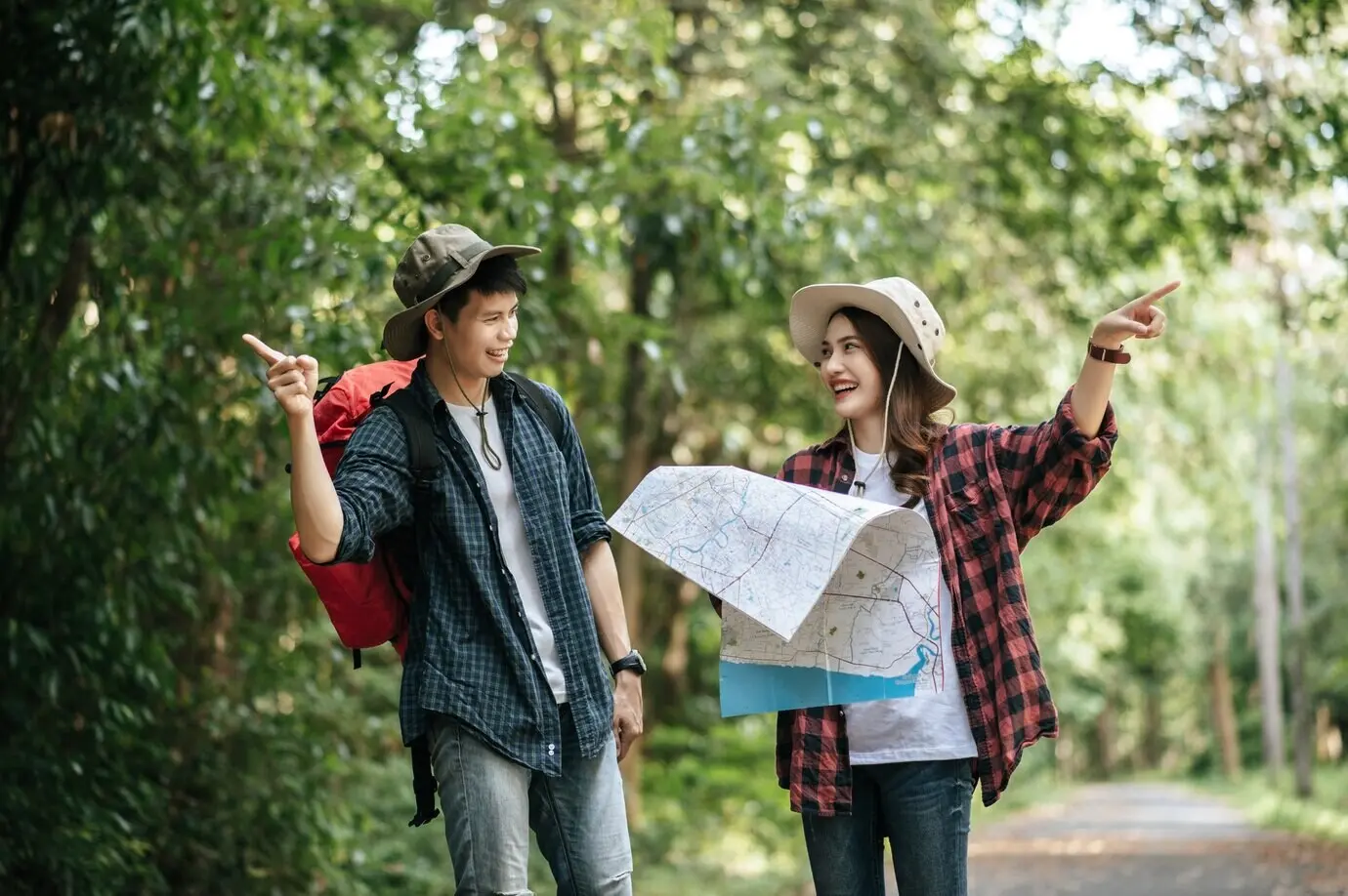 Portrait of a young, handsome Asian man with a backpack and a trekking hat and his pretty girlfriend, standing and checking direction on a paper map while walking on a forest trail; backpack travel concept.
